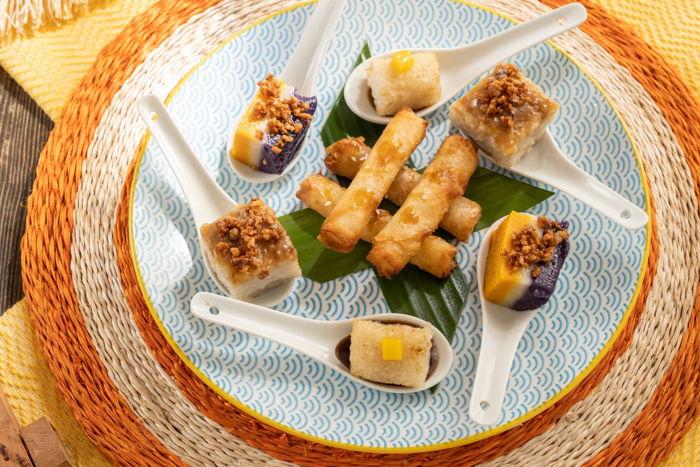 Dessert sampler of turon sticks, biko squares, ube and leche flan bites topped with latik in porcelain spoons on a pale blue patterned plate over a woven orange charger