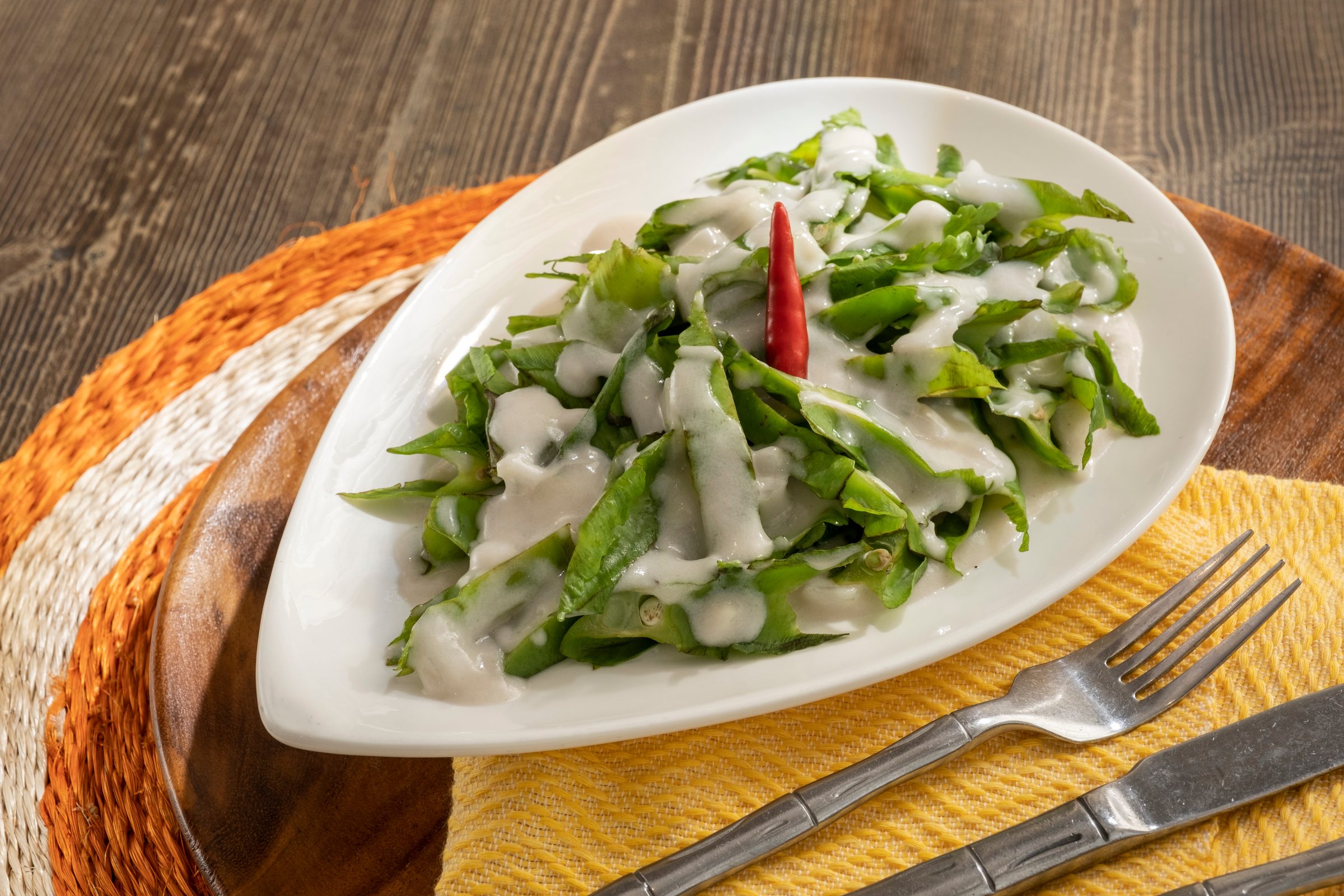 Sigarilyas, winged beans in creamy coconut sauce topped with a red chilli, on a white teardrop plate over a woven charger with a yellow cloth and cutlery