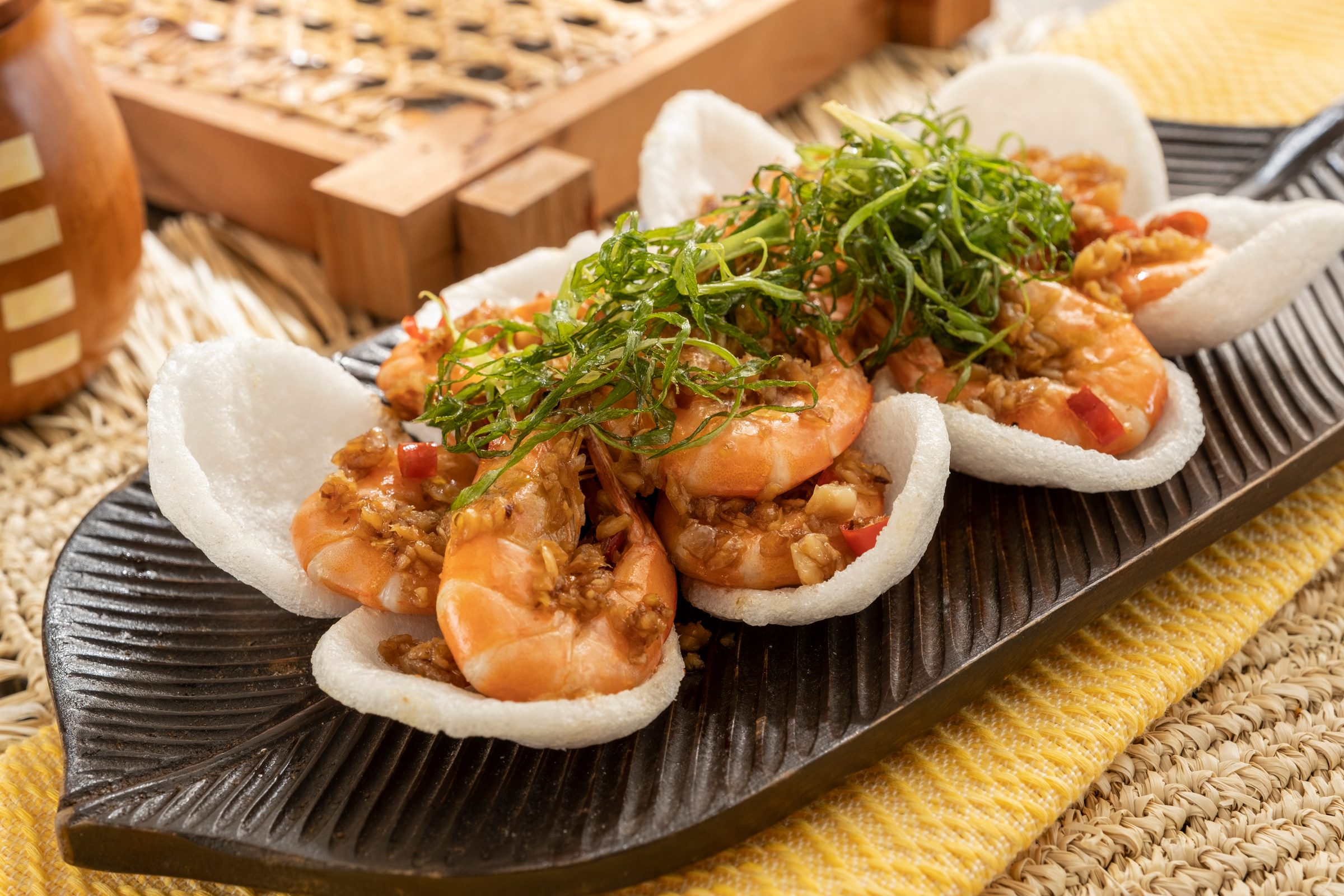 Stir-fried salted and spicy shrimp topped with crispy shredded herbs on prawn crackers on a dark leaf-shaped platter over a yellow cloth and woven mat
