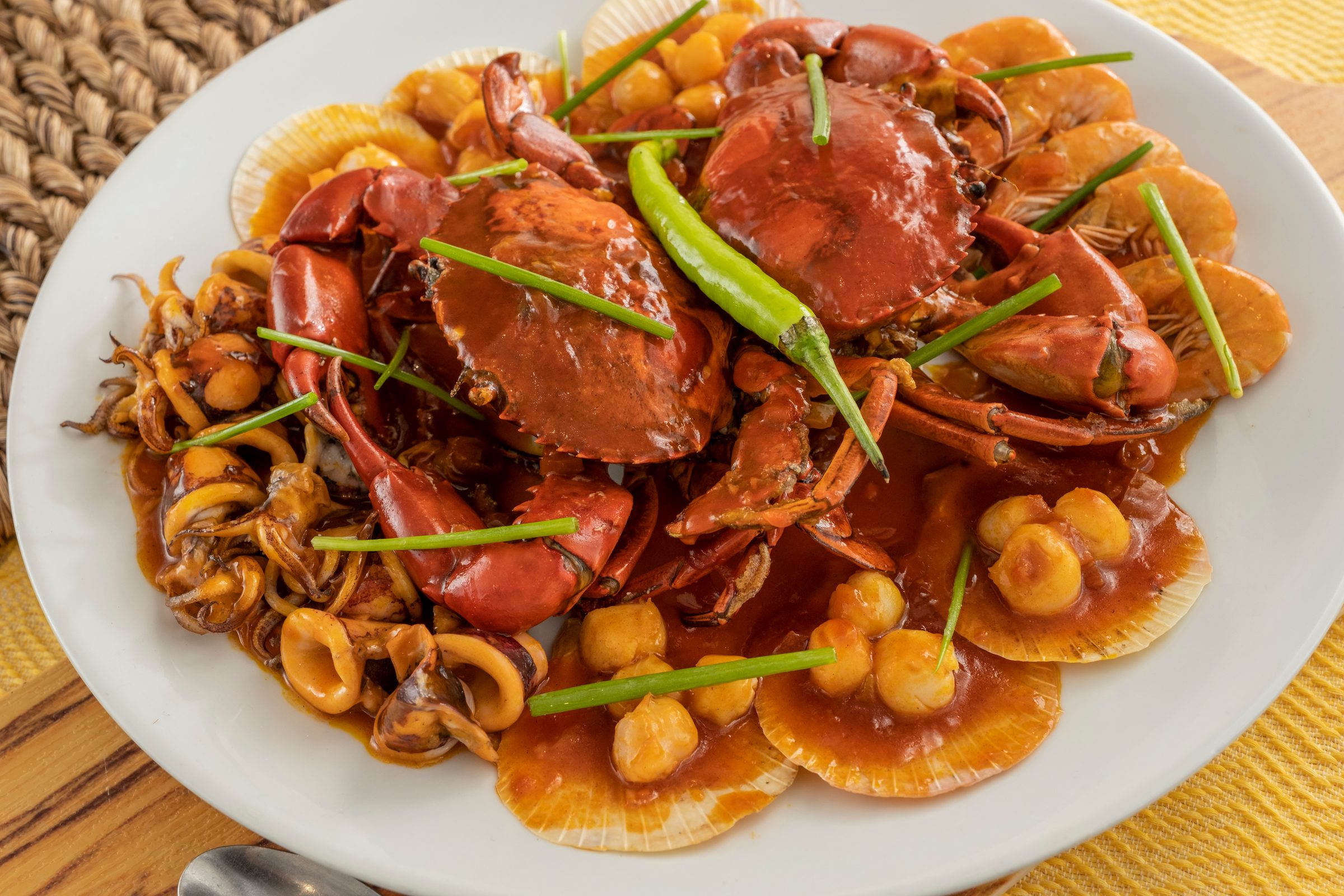 Crab and seafood mix in chilli sauce with whole crabs, squid rings and scallops, topped with a long green chilli, on a white plate over a yellow cloth and woven mat